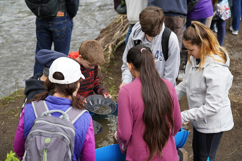 Six students gather around a water quality experiment. They each have a black bowl in their hands filled with water scooped from a half barrel. 
